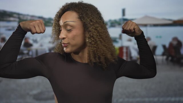 Transgender woman flexes biceps and raises fists in street with light blue background; empowerment pride.