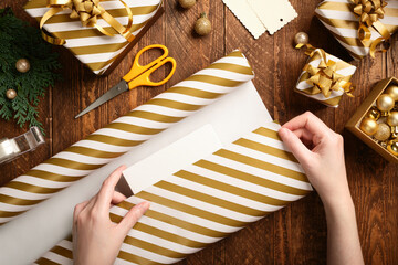Gift wrapping. Hands wrap a box in striped gold and white paper on a brown wooden table.
