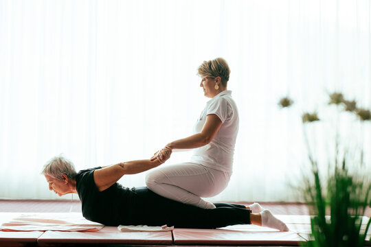 Professional female physiotherapist supporting an older woman during a therapeutic stretching exercise on a mat. Rehabilitation and physiotherapy concept focused on mobility, flexibility, recovery