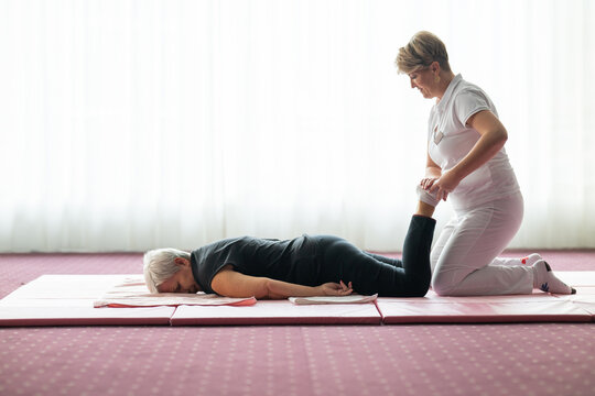 Professional female physiotherapist helping an older woman stretch her legs while lying face down on a mat. Rehabilitation and physiotherapy concept focusing on mobility recovery, flexibility, and