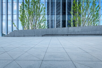 Empty square floor in front of a modern glass office building facade with green trees on a sunny day.