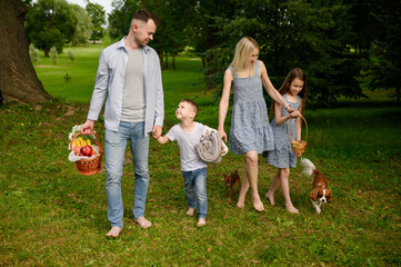Fototapeta premium Caucasian family is strolling through green park: father is carrying picnic basket, mother is holding daughter's hand, little boy is jumping nearby, and dogs running at feet. Family weekend