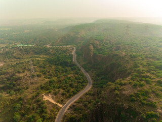 Aerial view of a solitary road winding through a dense, green forest, cutting a path through the rugged terrain under a hazy sky, Gurugram, Haryana, India.