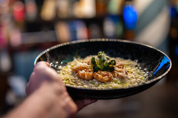 Delicious creamy shrimp and broccoli risotto in a black bowl held by a hand in a restaurant setting with a dark, blurred background