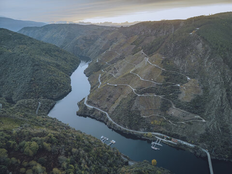 Aerial view of the Sil River cutting through the dramatic, vineyard-covered slopes of Ribeira Sacra, a tapestry of green and blue under a soft sky, Sober, Lugo, Spain.