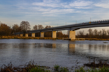 A wide view of a concrete bridge spanning a calm river on an autumn day, surrounded by dry grass, bare trees, and soft evening light.