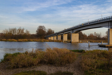 A wide view of a concrete bridge spanning a calm river on an autumn day, surrounded by dry grass, bare trees, and soft evening light.