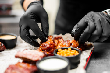 A close-up shot of saucy, hot chicken wings being arranged on parchment paper by a person wearing black gloves, alongside dipping sauces and sides.