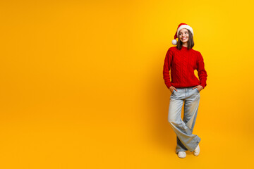 Fototapeta premium Playful Christmas vibe with a stylish young woman in a red sweater and Santa hat posing against a bright yellow background for holiday fashion and festive lifestyle themes