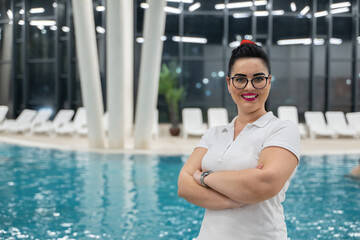 Portrait of a confident aquatic fitness instructor standing by the pool with senior women exercising in the background. Concept of health, wellness, active lifestyle, and rehabilitation training.