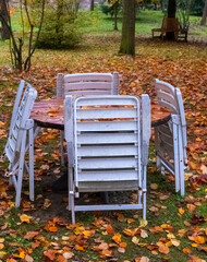 Chairs stacked around a weathered wooden table in an autumn park setting filled with fallen leaves