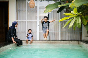A child jumps into the pool with excitement while his mother and younger brother sit by the edge enjoying family swim time.