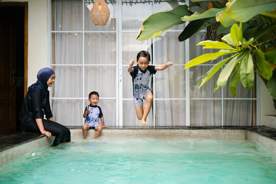 A child jumps into the pool with excitement while his mother and younger brother sit by the edge enjoying family swim time.