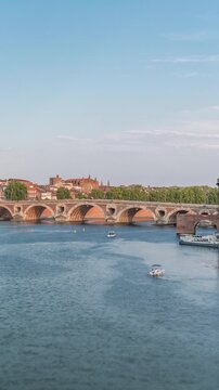 Garonne River and Pont Neuf timelapse with Port de la Daurade in downtown Toulouse, France. This Renaissance arch bridge reflects in the water under a blue sky with clouds. Waterfront with green trees