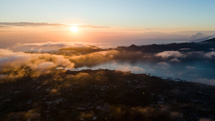 Aerial view of the sun kissing the clouds over the rugged terrain, painting the sky with hues of orange and gold, Mount Batur, Bali, Indonesia.