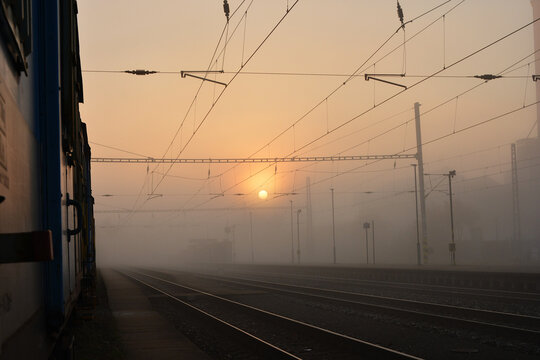 A train standing at a railway station platform during a foggy sunrise. Atmospheric morning scene with the sun glowing through the mist and overhead power lines