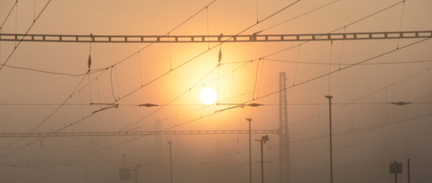A dramatic foggy sunrise over a railway line. Silhouettes of overhead power cables crisscross against the golden sun and orange sky in a misty atmosphere