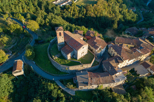 Aerial view of ancient stone church and fortress perched atop a hill, bathed in golden sunlight, surrounded by lush green forests, Trassilico, Tuscany, Italy.