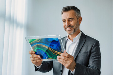 A smiling businessman in a suit holding a transparent tablet with a colorful abstract display, standing against a bright white background.