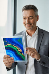 A smiling businessman in a gray suit holding and interacting with a tablet displaying an abstract blue and green marble design, photographed in a bright office setting.