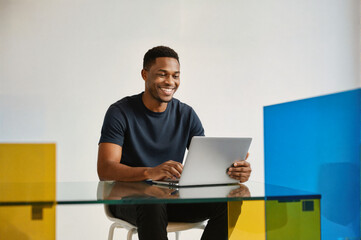 A smiling young man in a black t-shirt working on a laptop at a glass desk in a modern office with colorful geometric panels.