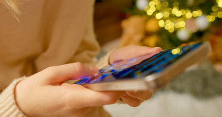 Woman engaging with smartphone near illuminated Christmas tree, enjoying festive connectivity and seasonal digital moments at home