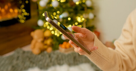 Young woman relaxing near illuminated Christmas tree, browsing smartphone while sitting by warm fireplace during festive winter holiday season