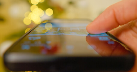 Close up finger scrolling on smartphone screen with Christmas lights bokeh effect in background