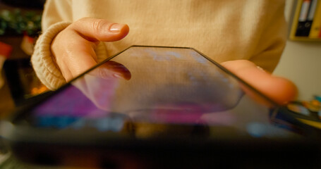 Young woman leisurely scrolling through social media news feed on smartphone while sitting comfortably near soft christmas lights with warm bokeh background