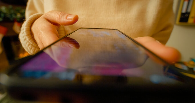 Young woman leisurely scrolling through social media news feed on smartphone while sitting comfortably near soft christmas lights with warm bokeh background