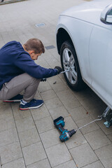 Man changing car tire using electric drill and jack on city sidewalk. DIY auto repair, maintenance, seasonal tire change, mechanical work, vehicle service, urban life.