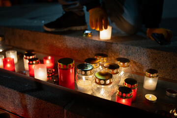 Warm memorial candles arranged on stone steps with a gloved hand placing a glowing lantern among assorted glass jars, highlighting a detailed nighttime tribute scene with soft reflections