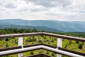 Fototapeta premium Scenic mountain panorama from wooden observation deck with railing, overlooking conifer forest and distant hills under cloudy summer sky, empty viewpoint with copy space for travel, tourism concepts