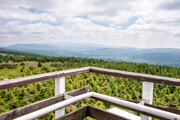 Fototapeta premium Scenic mountain panorama from wooden observation deck with railing, overlooking conifer forest and distant hills under cloudy summer sky, empty viewpoint with copy space for travel, tourism concepts