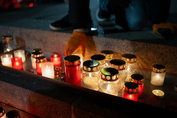 Multiple warm lanterns placed in a neat row across stone steps as a gloved hand lowers a candle into position, emphasizing coordinated memorial lighting