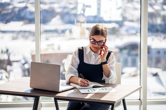 Professional woman engaged in a business call while analyzing financial reports in a modern office overlooking a snowy cityscape