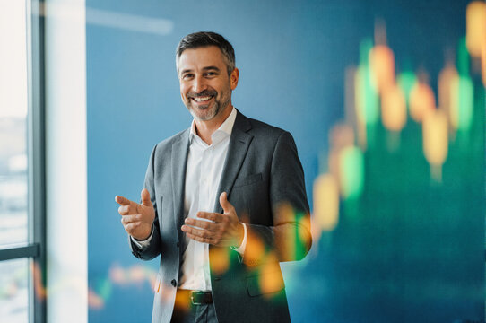 A smiling businessman in a suit stands in an office, gesturing with his hands as he presents financial data displayed on a screen behind him.