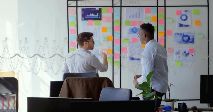 Seated man starting business plan, pointing at glass charts board, peer using animated overlay