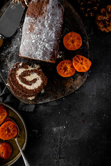 Homemade chocolate roulade filled with vanilla cream, dusted with icing sugar, and served alongside glossy candied orange slices. Presented on a rustic wooden board with pinecones and dark textured ba