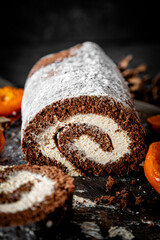 Homemade chocolate roulade filled with vanilla cream, dusted with icing sugar, and served alongside glossy candied orange slices. Presented on a rustic wooden board with pinecones and dark textured ba