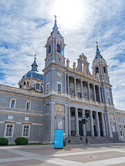 The Almudena Cathedral in Madrid, showcasing its neo-Romanesque exterior, with twin bell towers, a central dome, and a large stone staircase under a dramatic blue and white sky.