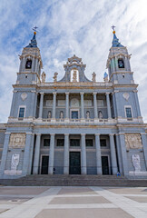 The Almudena Cathedral in Madrid, showcasing its neo-Romanesque exterior, with twin bell towers, a central dome, and a large stone staircase under a dramatic blue and white sky.