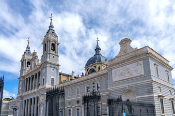 The Almudena Cathedral in Madrid, showcasing its neo-Romanesque exterior, with twin bell towers, a central dome, and a large stone staircase under a dramatic blue and white sky.