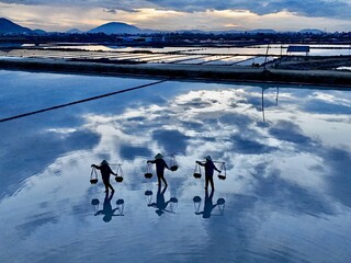 Ninh Hoa, Vietnam - 25 June 2025: Aerial view of workers harvesting salt on reflective salt ponds, their silhouettes mirrored in the tranquil water, against a backdrop of distant mountains.