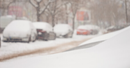 Gentle snow falls over a busy city landscape, blanketing cars and streets in a serene winter scene. The quiet charm of the snow transforms the familiar surroundings.