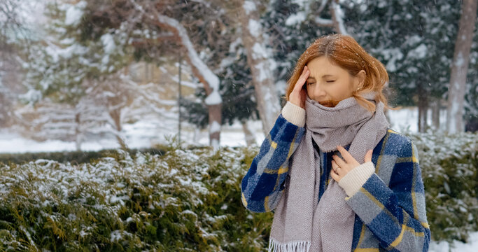 In a winter park, a redhead woman walks while snow falls gently around her. She gently holds her scarf, feeling discomfort from a headache amid the serene snowfall.