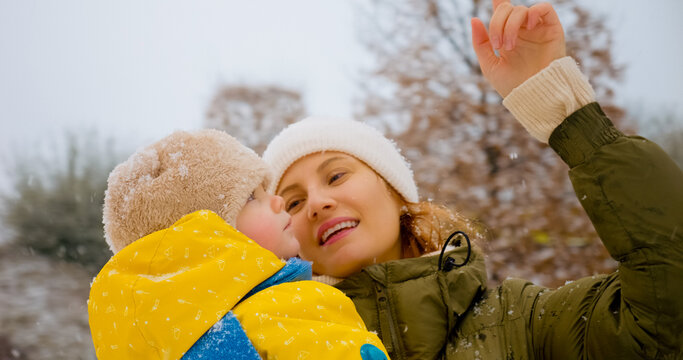A mother and her child revel in the snow, laughing and pointing at falling flakes while enjoying a joyful moment together in the winter park setting.