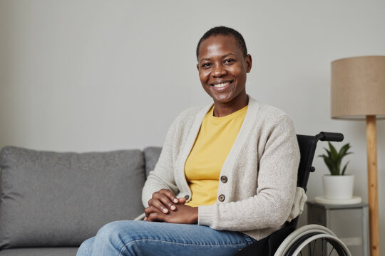 Portrait of young adult Black woman sitting in wheelchair with disability smiling at camera hands resting on lap modern living room background visible behind subject