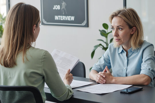 Caucasian middle aged woman sitting at desk facing young Caucasian woman holding documents, both engaged in job interview conversation in modern office setting - Powered by Adobe