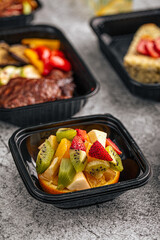 Colorful fresh fruit salad with kiwi, orange, apple and strawberries served in a black takeaway container on a textured background